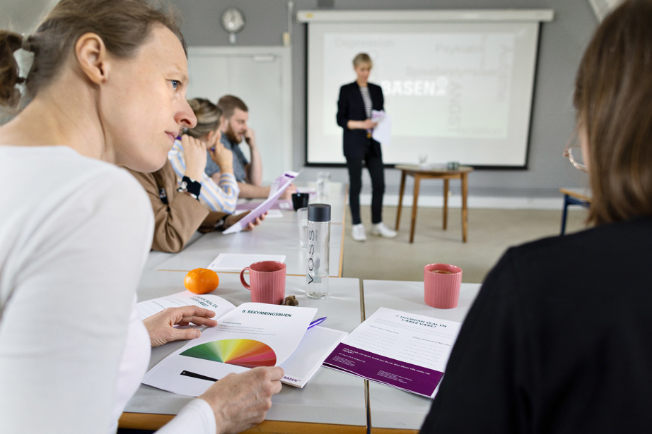Kvinde ved tavle i undervisningslokale med fire deltagere. To kvinder sidder og snakker sammen i forgrunden.